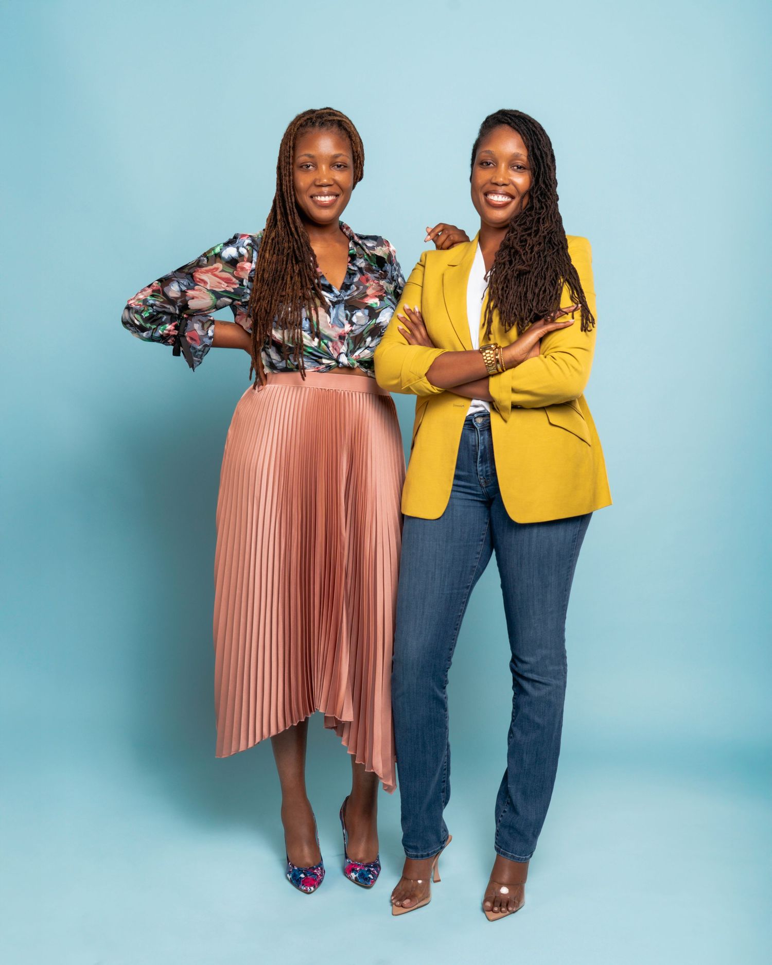 Two people wearing contrasting outfits of floral print and yellow stand together against a blue background.