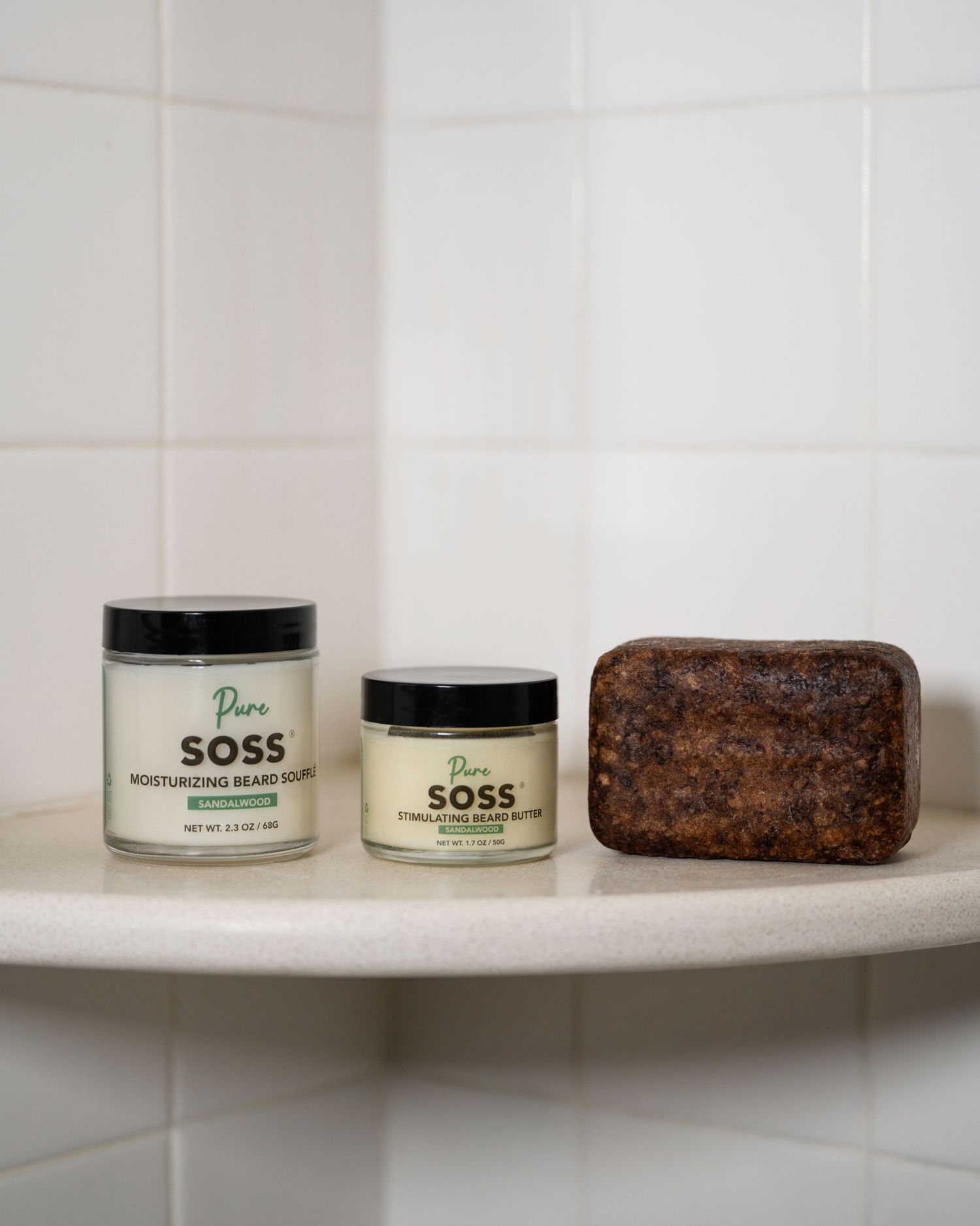 Two SOSS skincare product jars next to African black soap on white tile shelf.