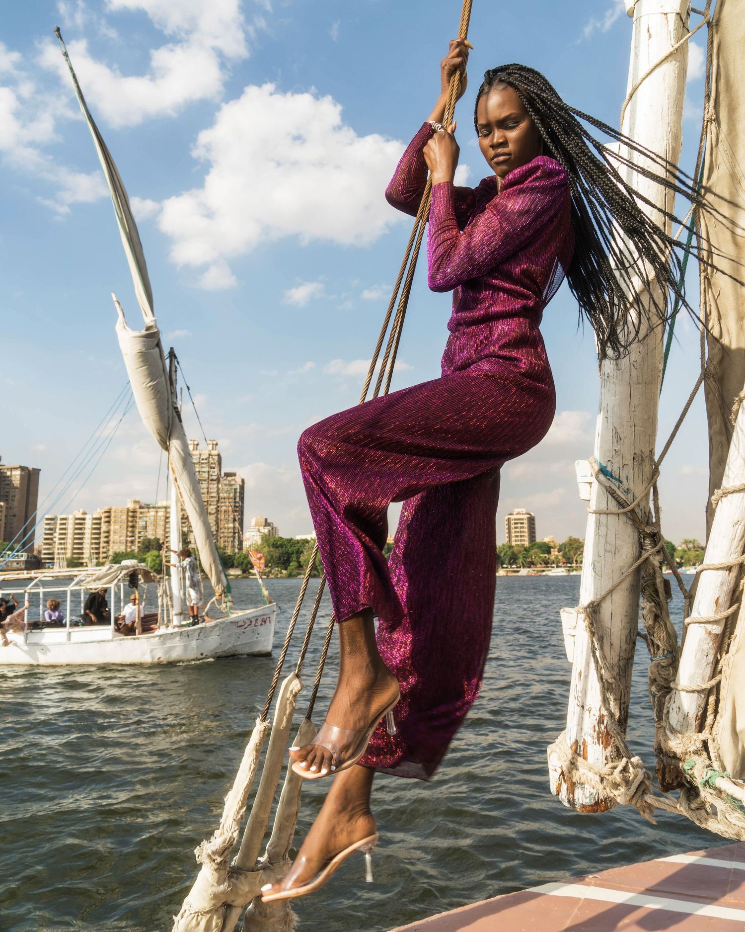 Fashion shoot on sailboat featuring purple jumpsuit against urban waterfront.