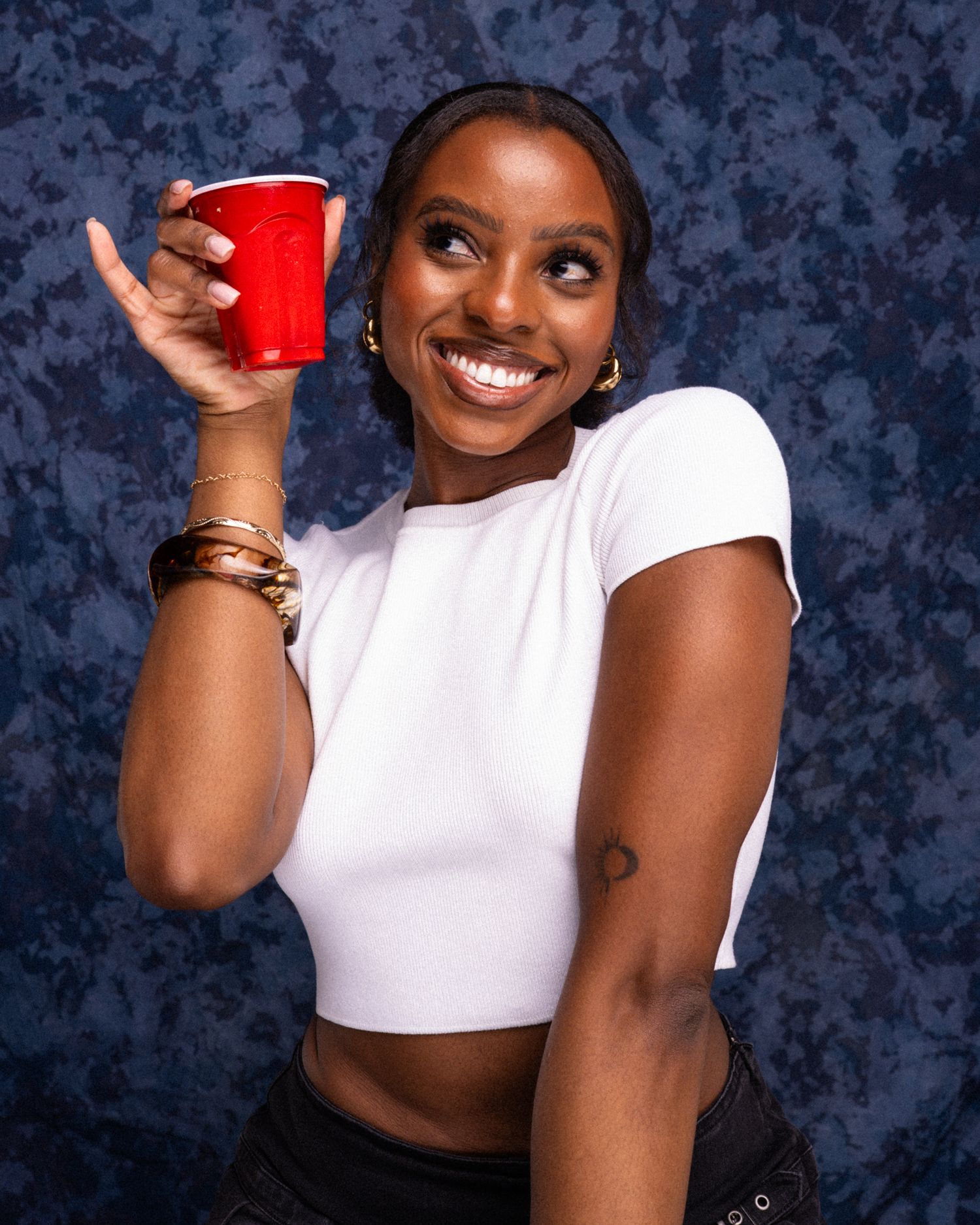 Smiling young woman in white crop top holding red cup, posing against blue mottled background.