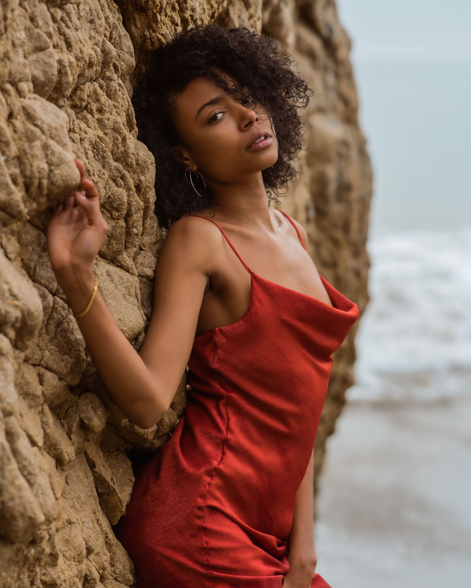A young woman in a red satin slip dress leans against a rocky cliff at the beach, posing gracefully.