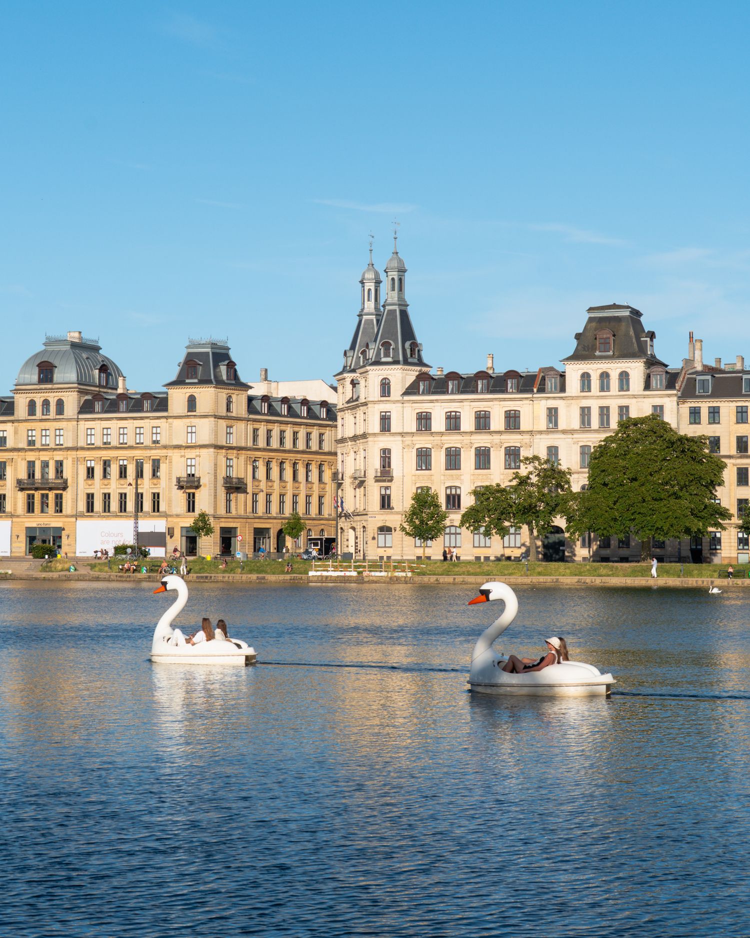 Swan-shaped paddle boats float in front of a grand palace on a serene lake.