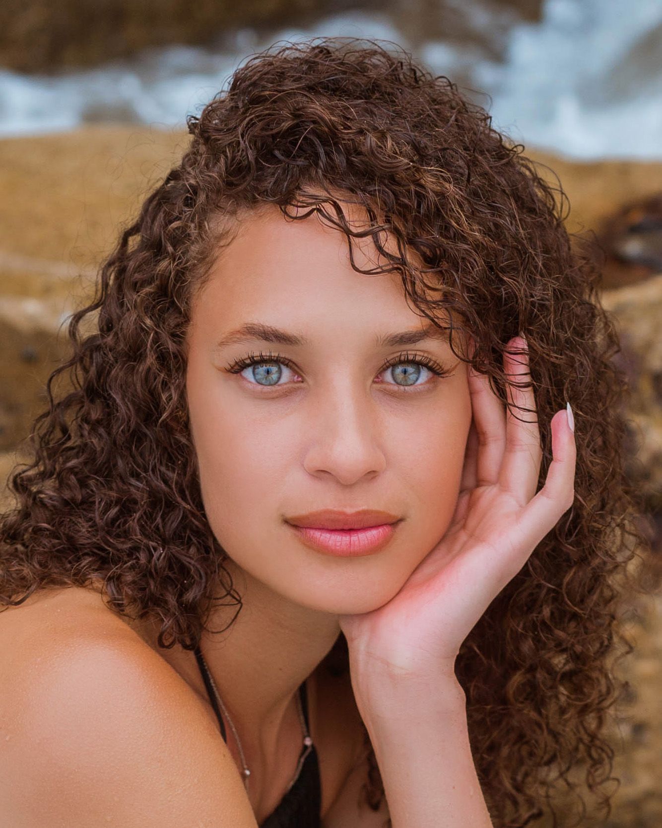A close-up portrait of a beautiful girl with curly brown hair, bright blue eyes and natural makeup against a soft beach background.