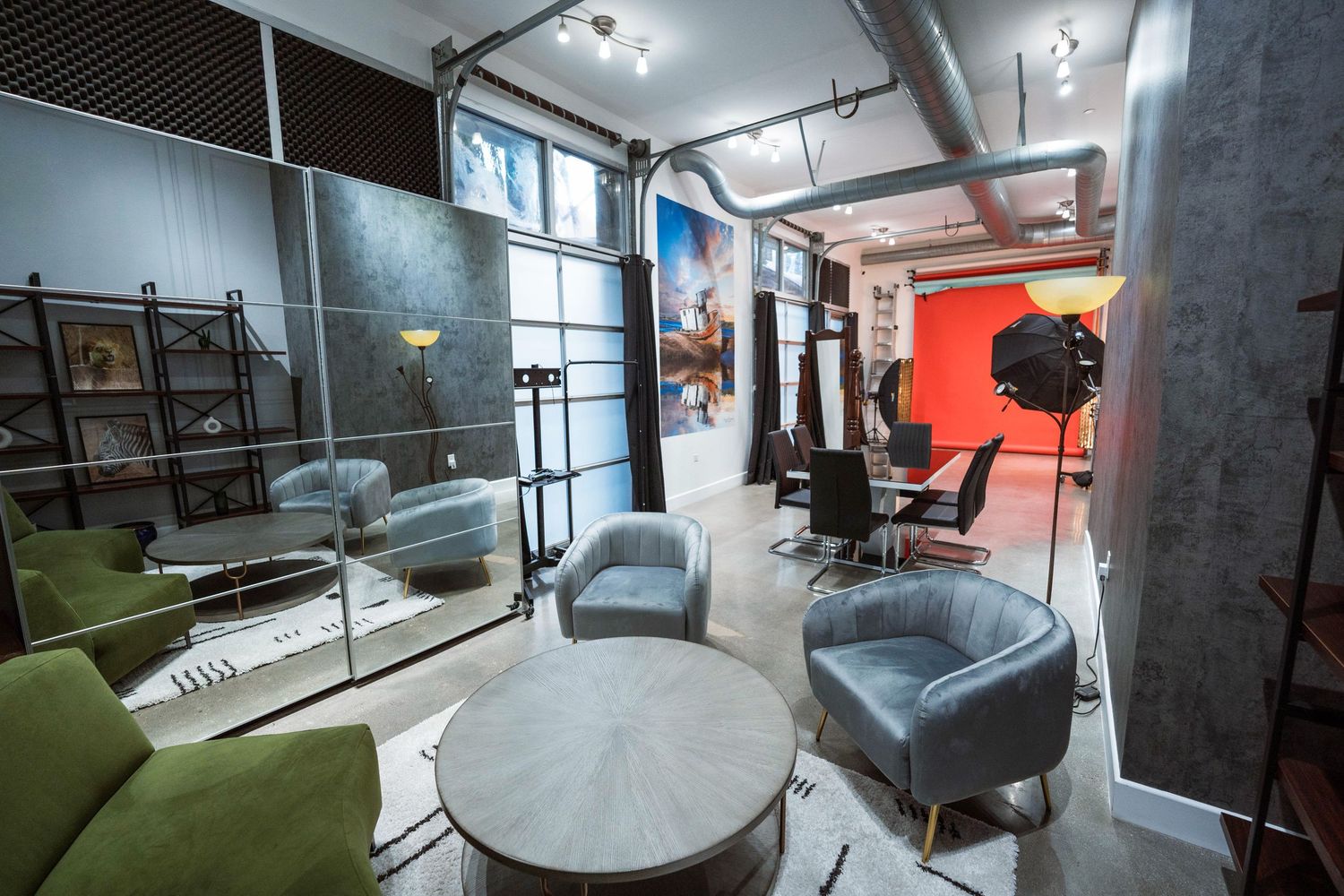 Modern studio lounge with gray armchairs, green sofa, round coffee table, large mirror, and professional photography lighting equipment.