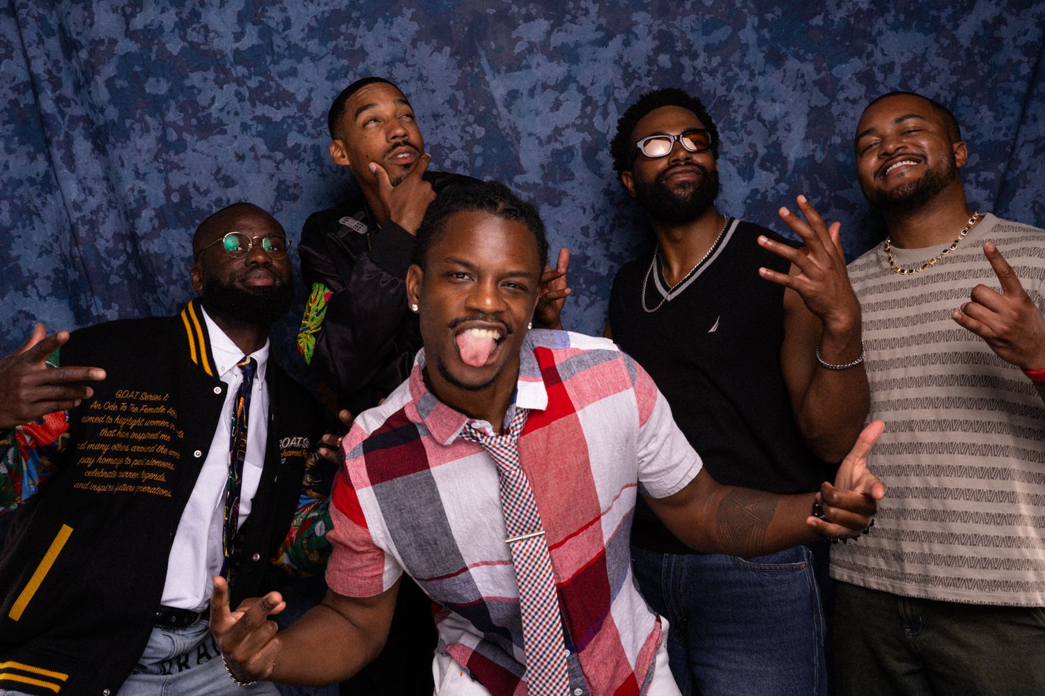 Five young men posing energetically in front of a blue backdrop, making fun gestures and smiling.