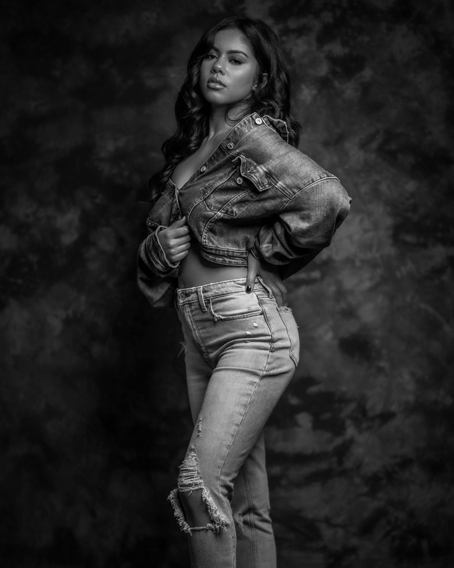 Black and white studio portrait of a woman in ripped jeans and denim jacket posing confidently against dark background.