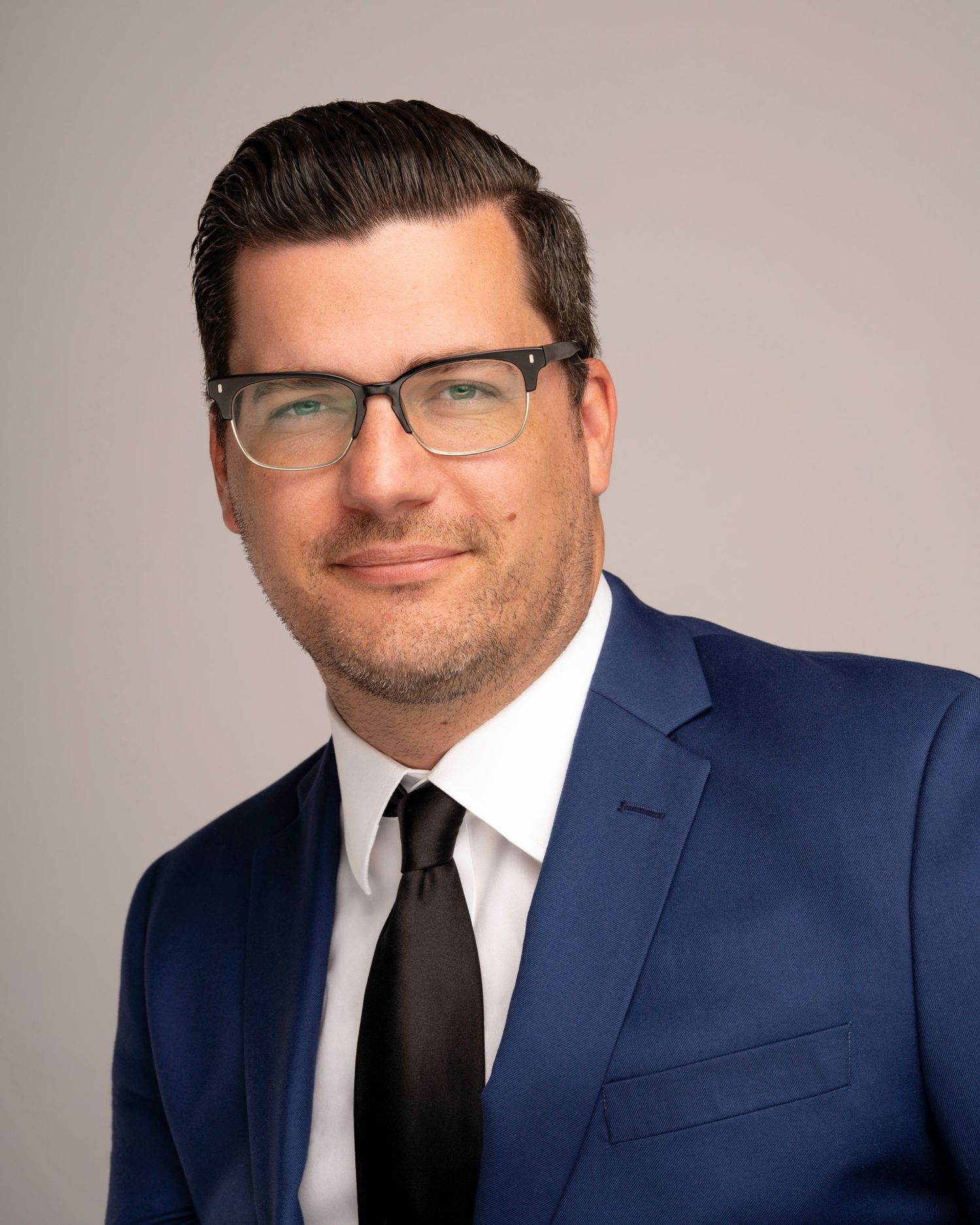 Professional headshot of a person wearing glasses, a navy suit, and black tie against a neutral background.