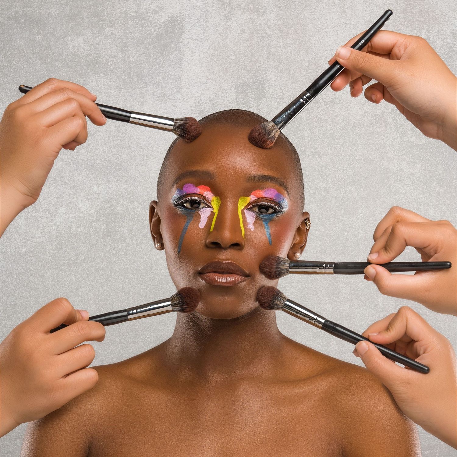 Beauty editorial close-up of a woman’s face being styled with multiple makeup brushes, captured against a neutral gray background.