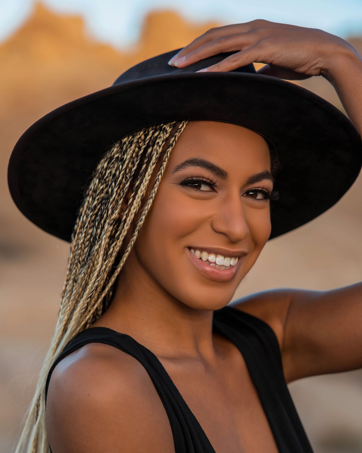 Smiling woman in black tank top and wide brim hat with long blonde braids poses outdoors at sunset.