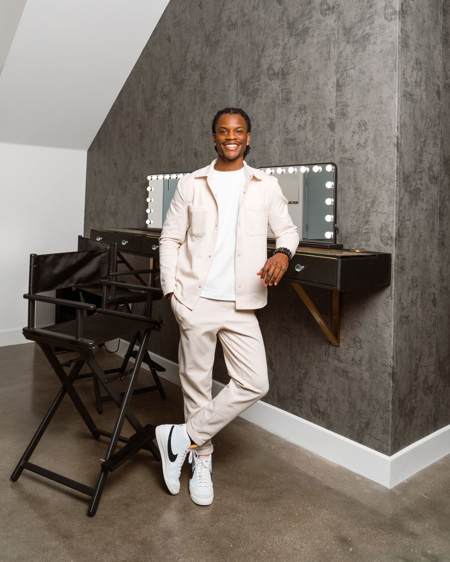 A person in a crisp white outfit sits on a modern black director's chair against a textured grey wall.