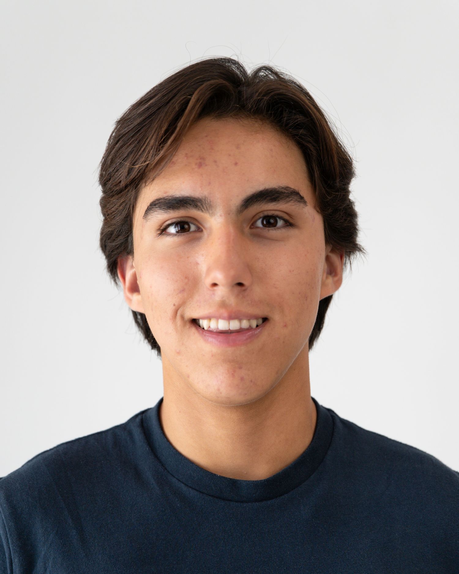 Young man with dark hair smiling, wearing a navy blue crew neck shirt against a light gray background.