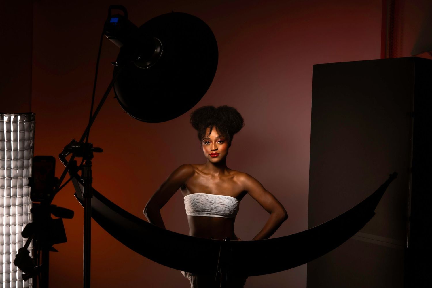 Young Black woman in white strapless top poses confidently in professional photo studio with lighting equipment.