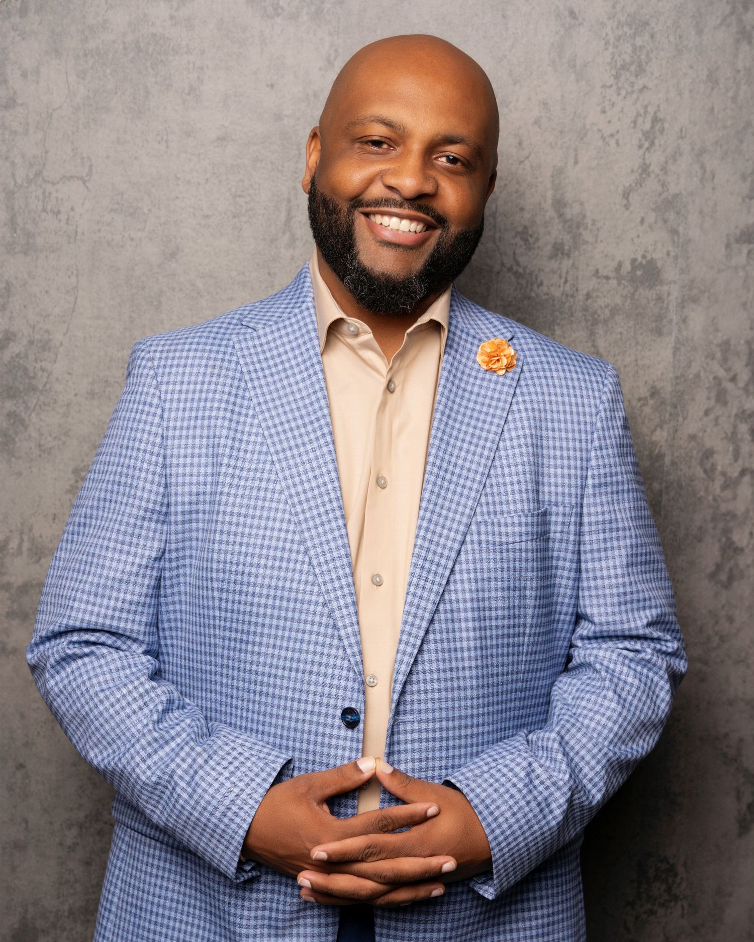 A person in a light blue checkered blazer and peach shirt poses against a textured gray backdrop in a professional headshot series.