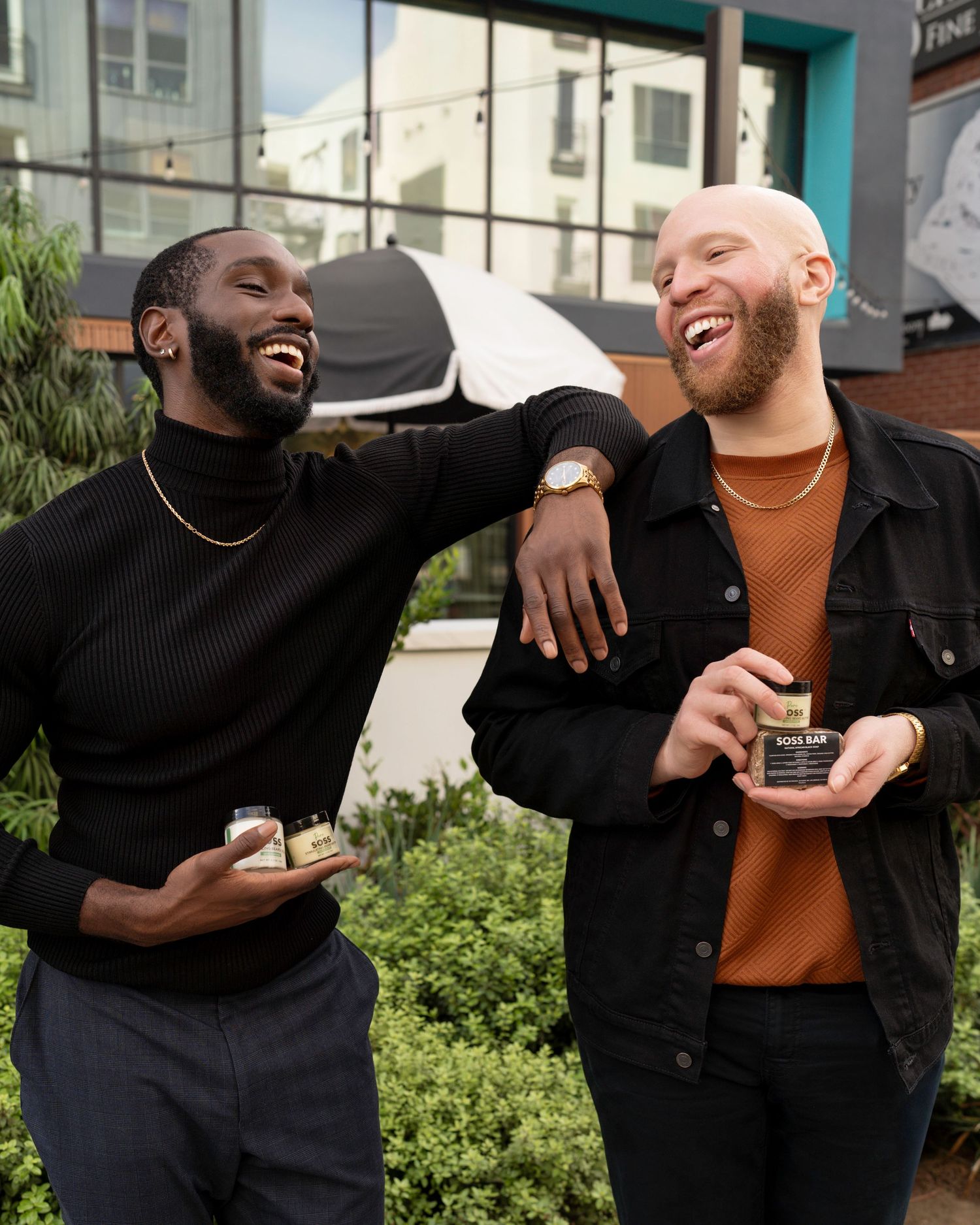 Two stylish men laughing and holding skincare products during a lifestyle brand photoshoot in an outdoor urban setting.