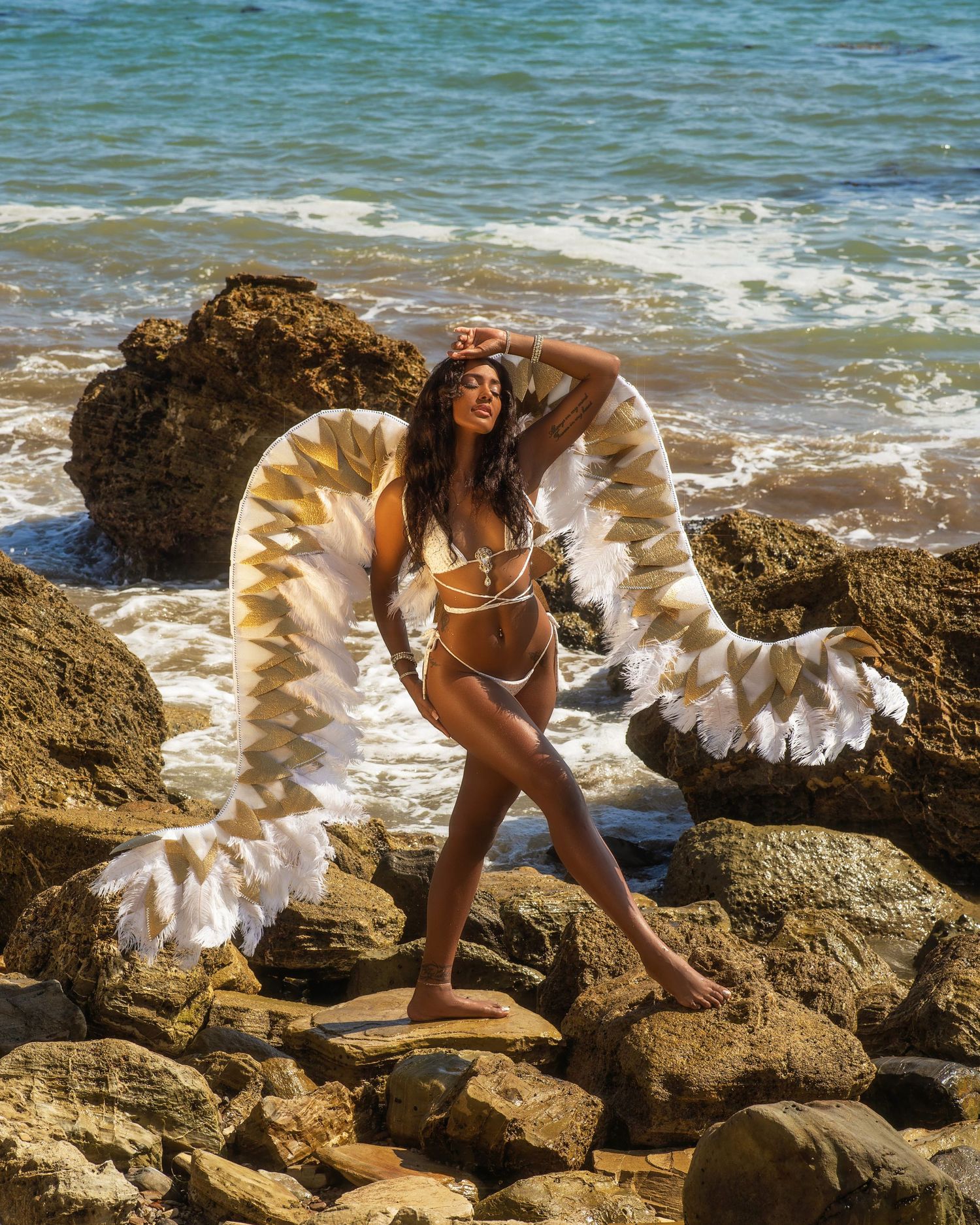 Model poses with angel wings on rocky beach shore in swimwear.