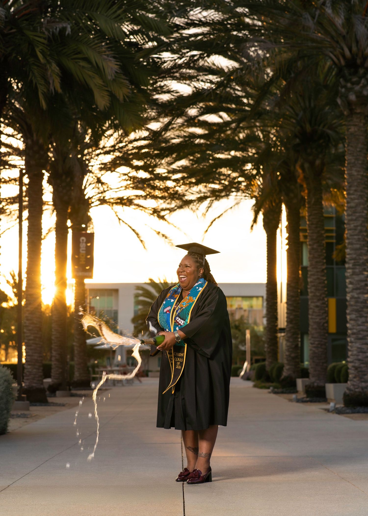 Graduate in cap and gown poses among palm trees at golden hour sunset on college campus.