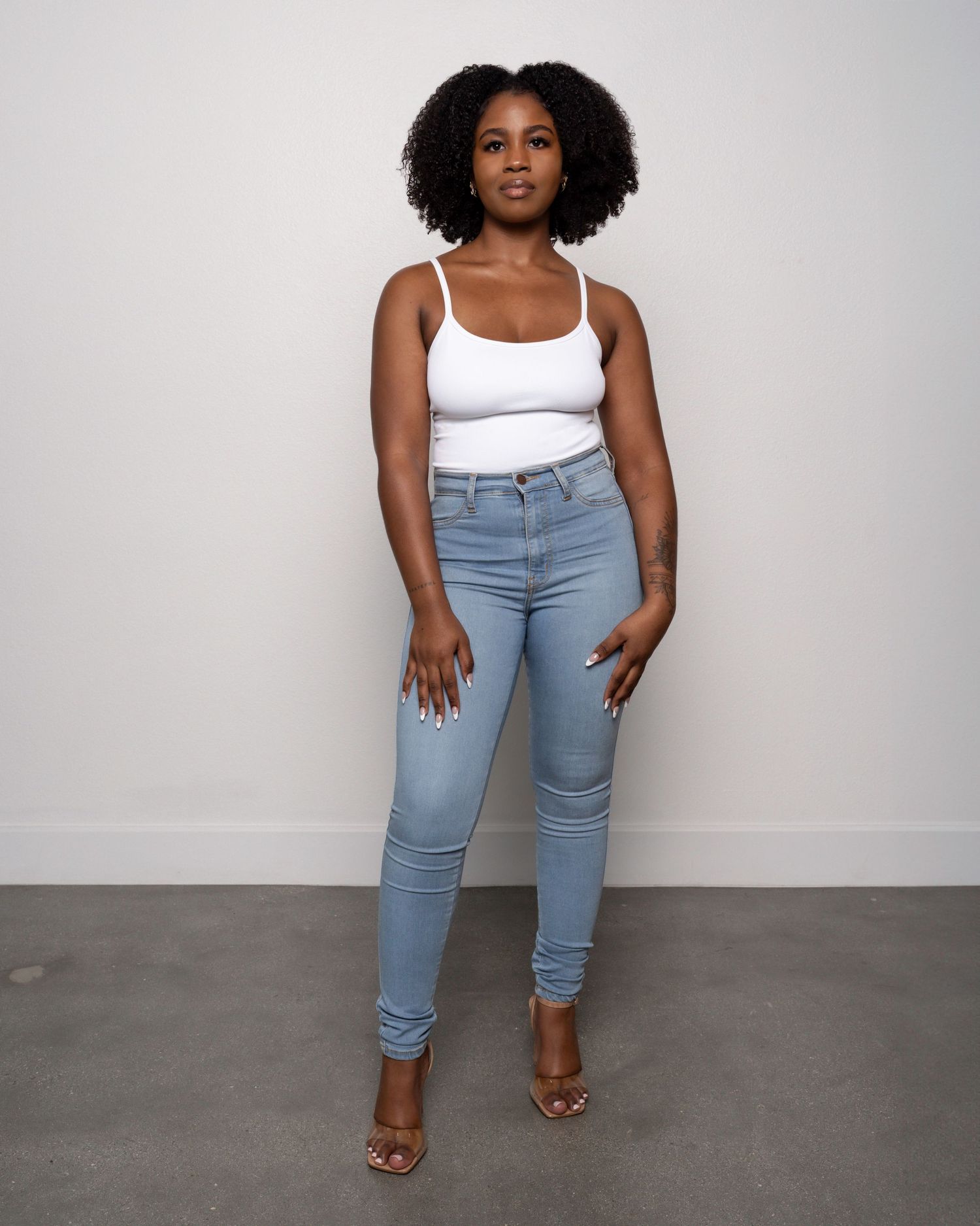 Woman wearing white tank top and light blue skinny jeans with clear heeled sandals, posing against white wall.