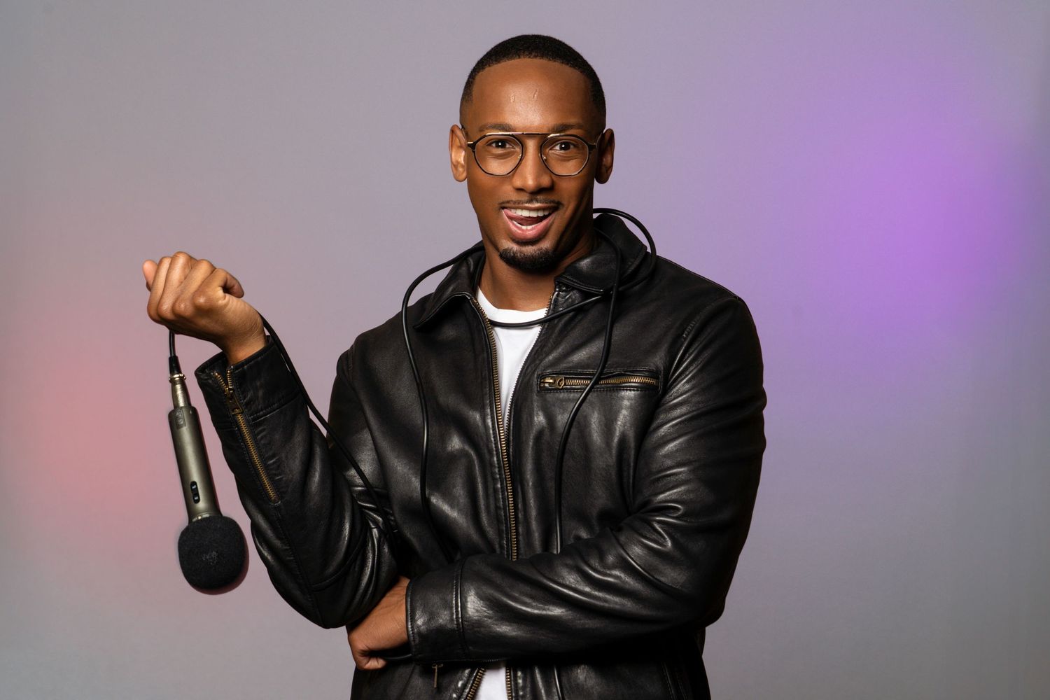 Person in leather jacket holding microphone against purple gradient background.