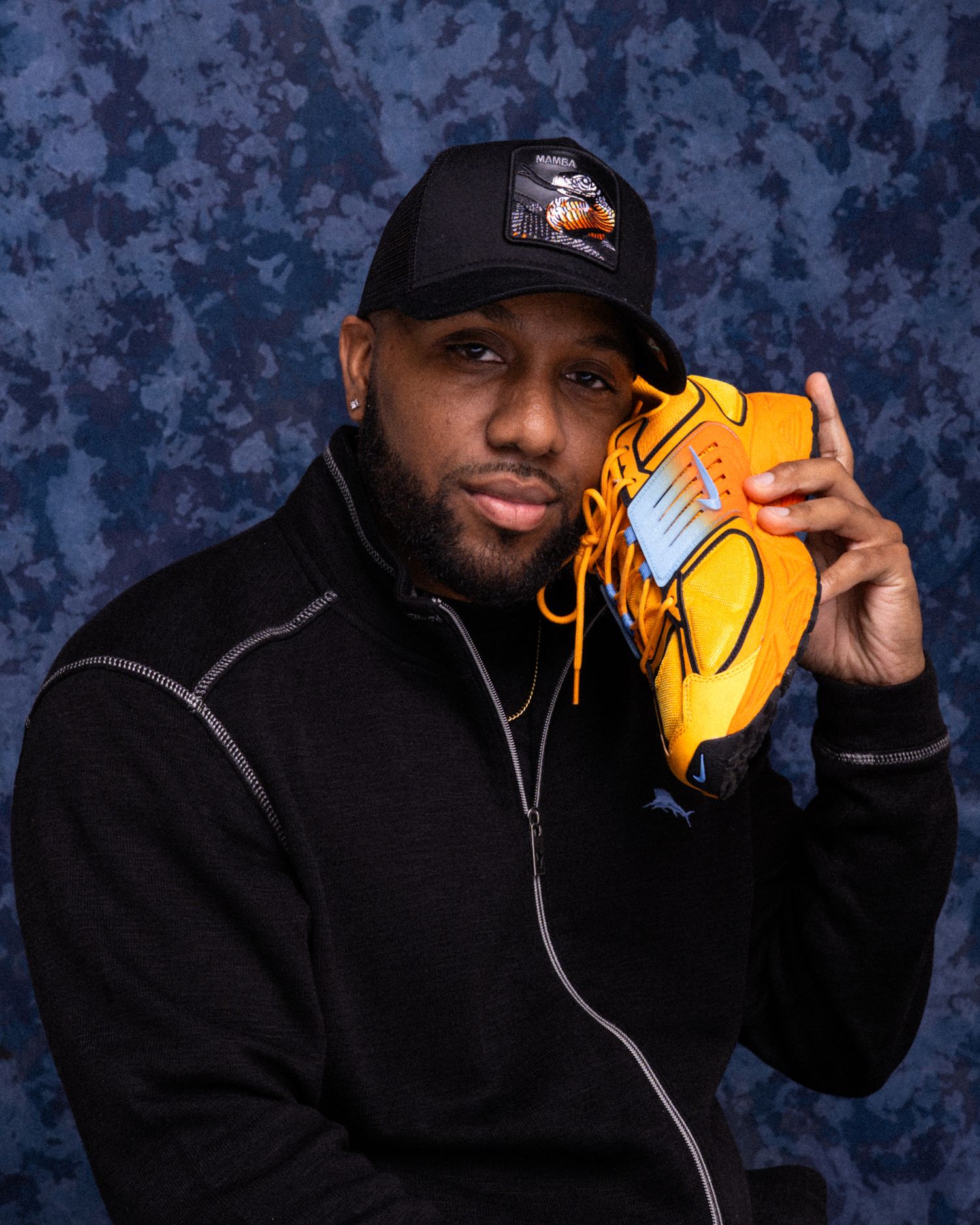 Man in black jacket and cap holds orange baseball glove near face against blue backdrop.