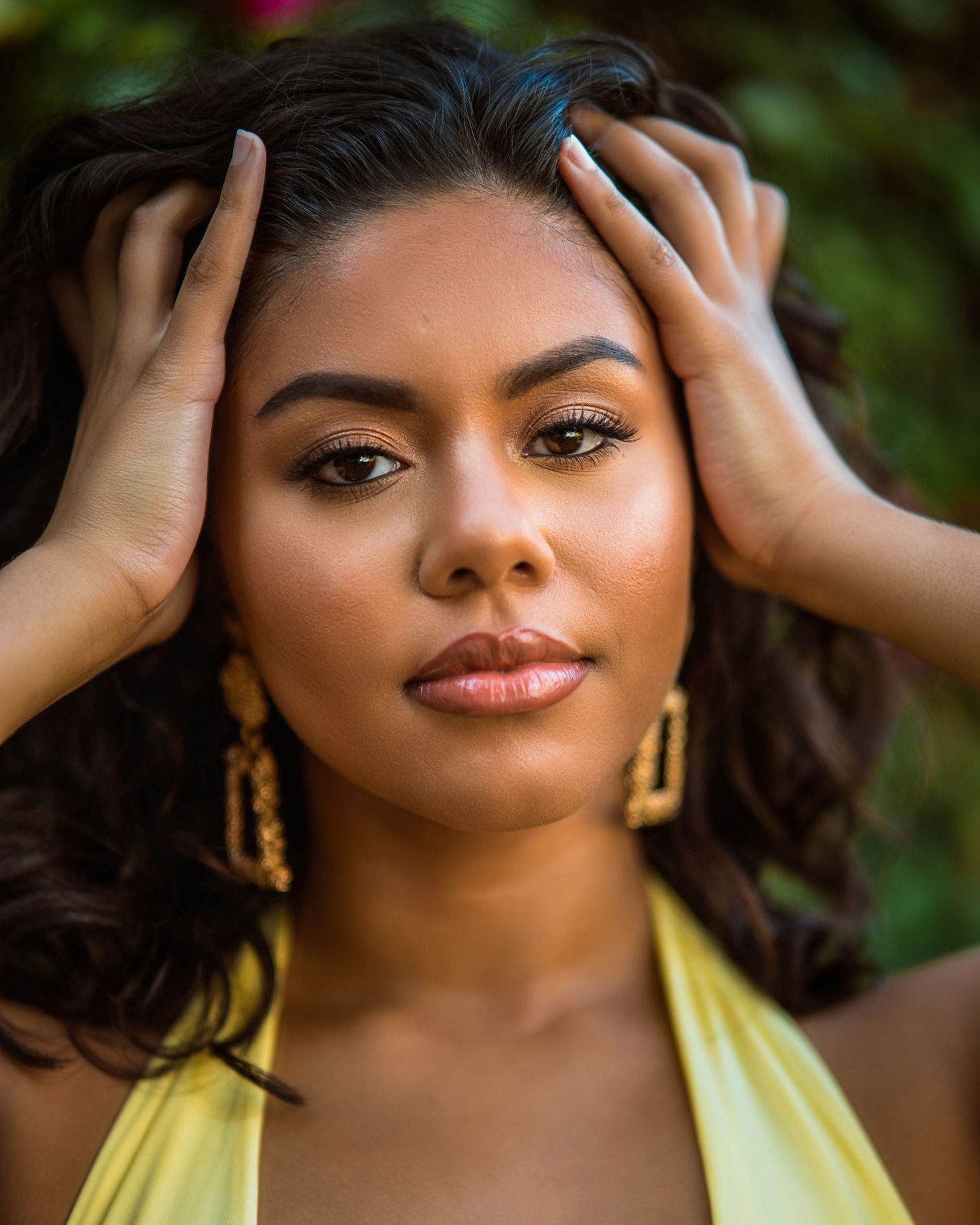 Young woman with gold earrings and yellow top poses with hands in dark wavy hair against green foliage backdrop.
