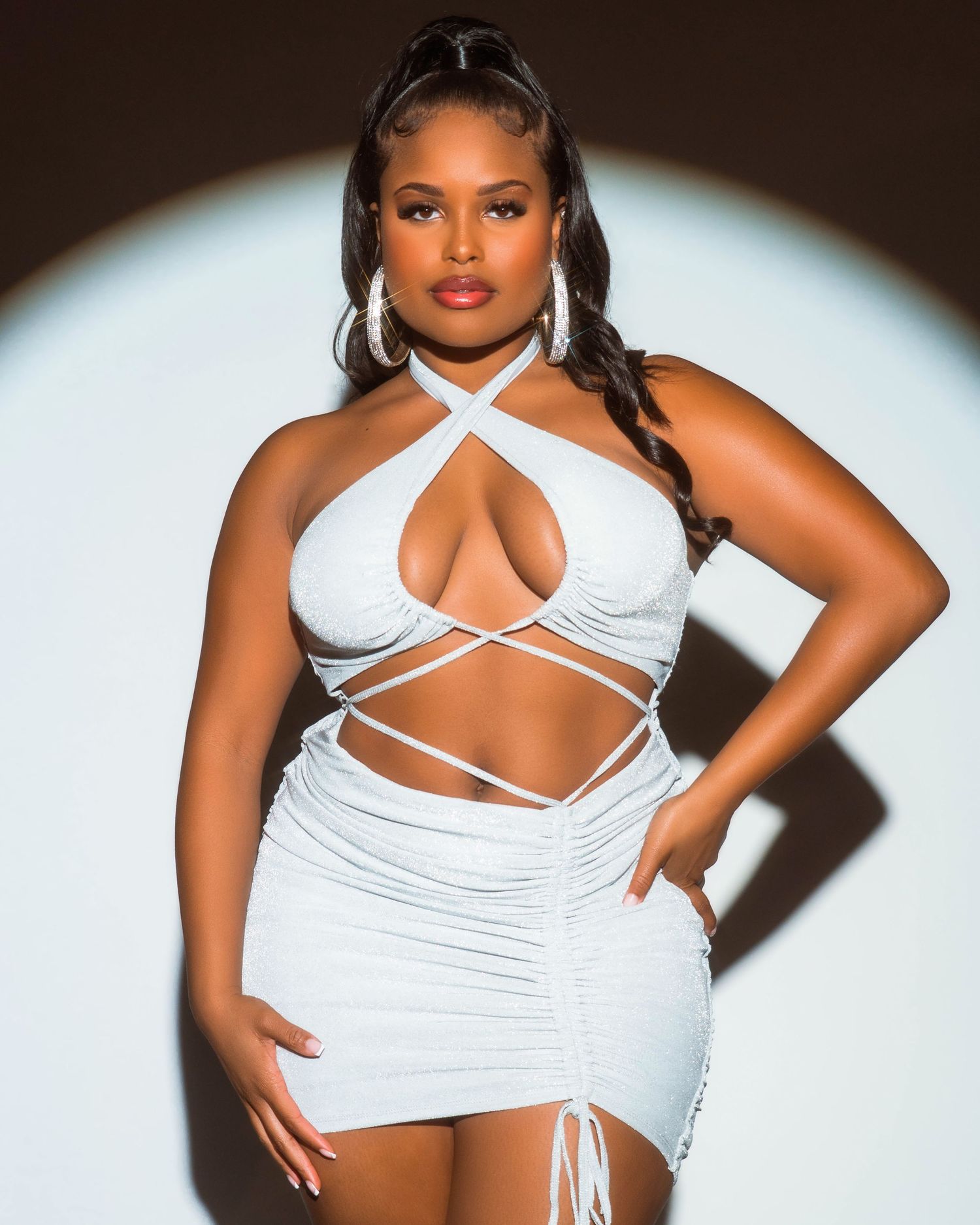 Fashion model in white strappy top poses against dark background in glamour photoshoot series.