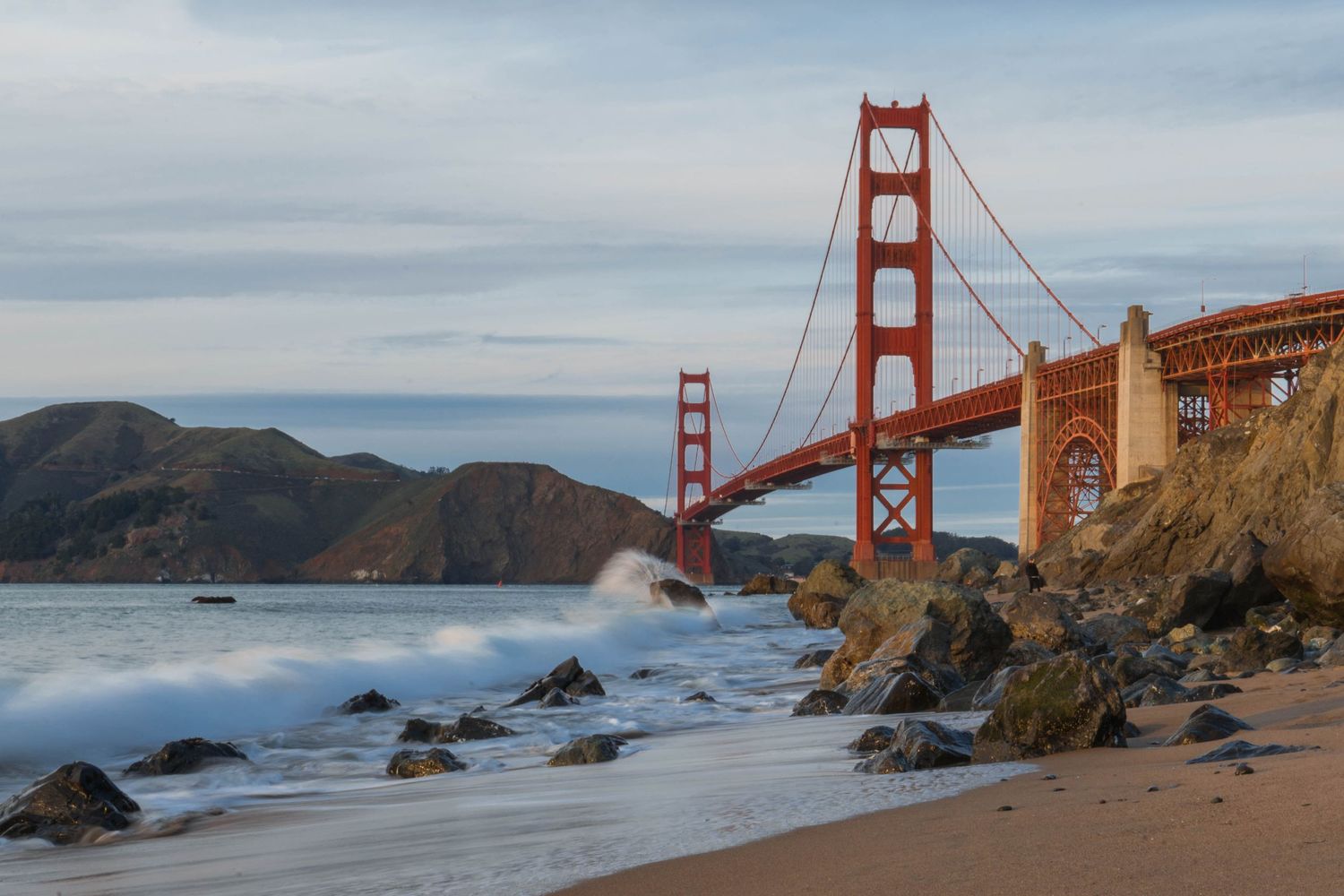 The iconic Golden Gate Bridge spans over San Francisco Bay with rocky beach and misty coastline at sunset.
