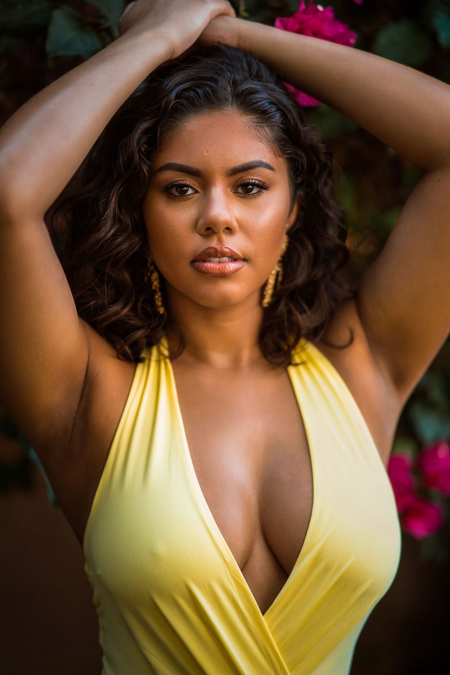 A young woman in a yellow deep-V swimsuit poses with hands raised, wearing gold earrings, with pink flowers in background.