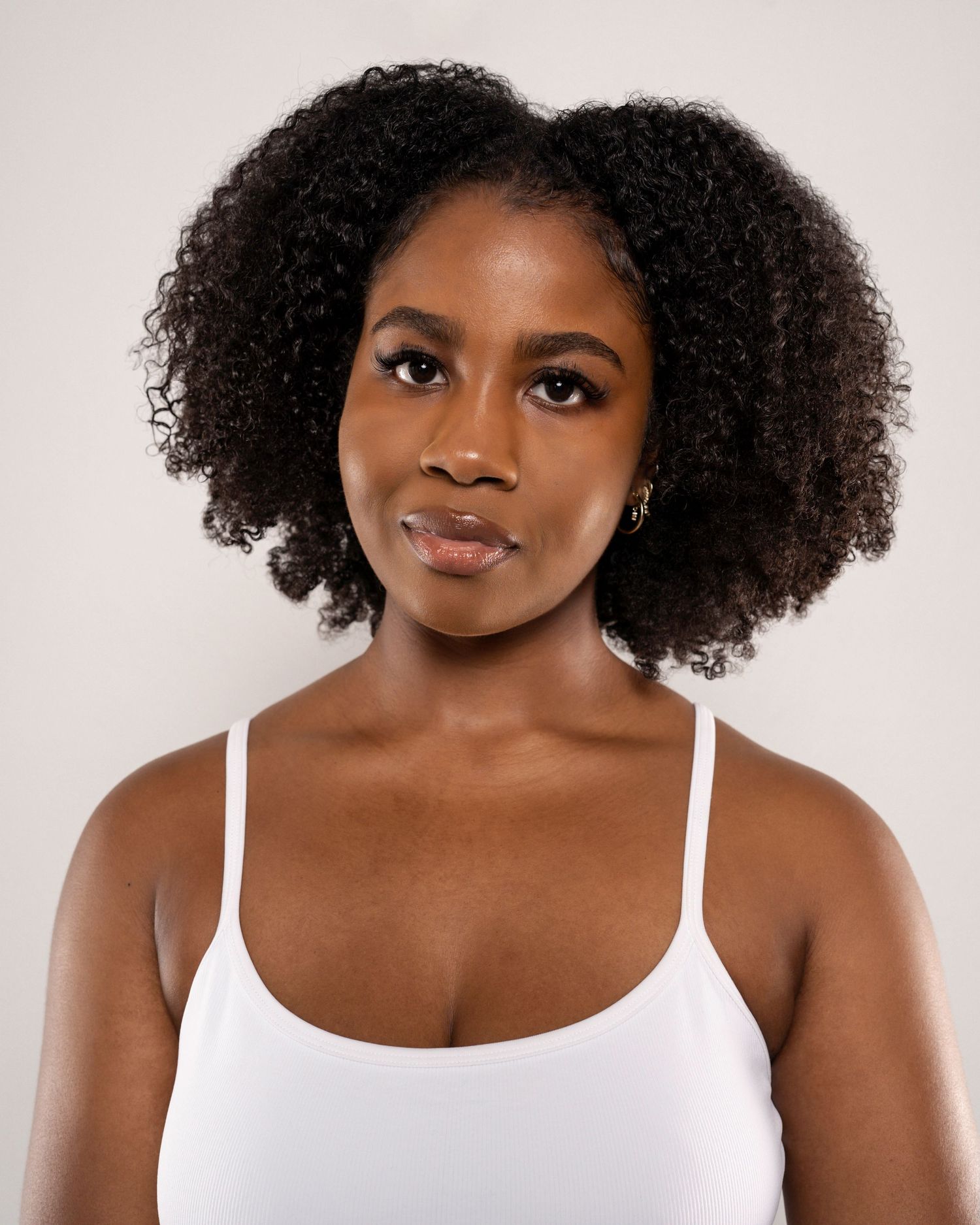 Young Black woman with natural curly hair wearing a white tank top poses against a light background.
