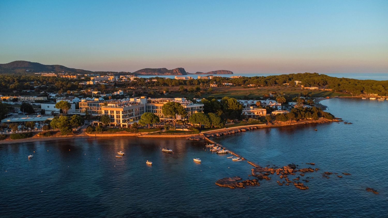 Aerial photograph of a coastal resort and marina in Ibiza at golden hour, featuring calm water, boats, and scenic shoreline architecture.