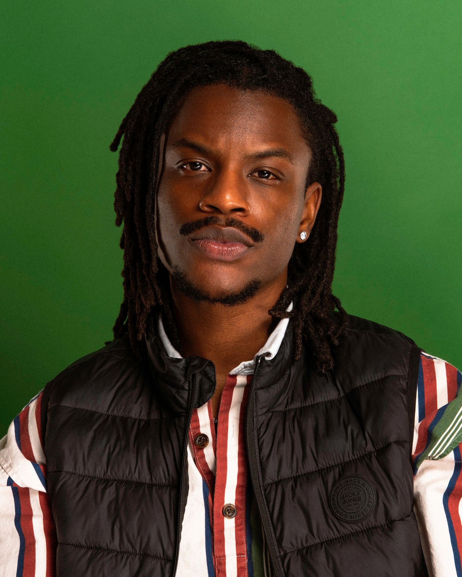 Person wearing a black puffer jacket and striped shirt smiles while posing against a green background.