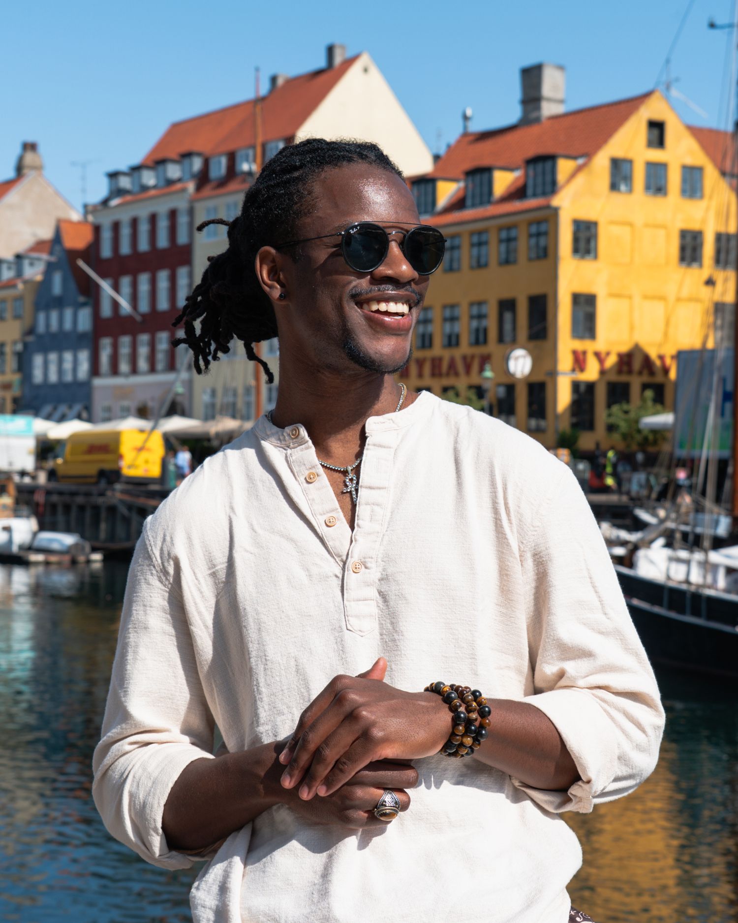 Handsome young man in linen shirt and sunglasses smiling by colorful waterfront buildings and boats in Nyhavn, Copenhagen harbor.