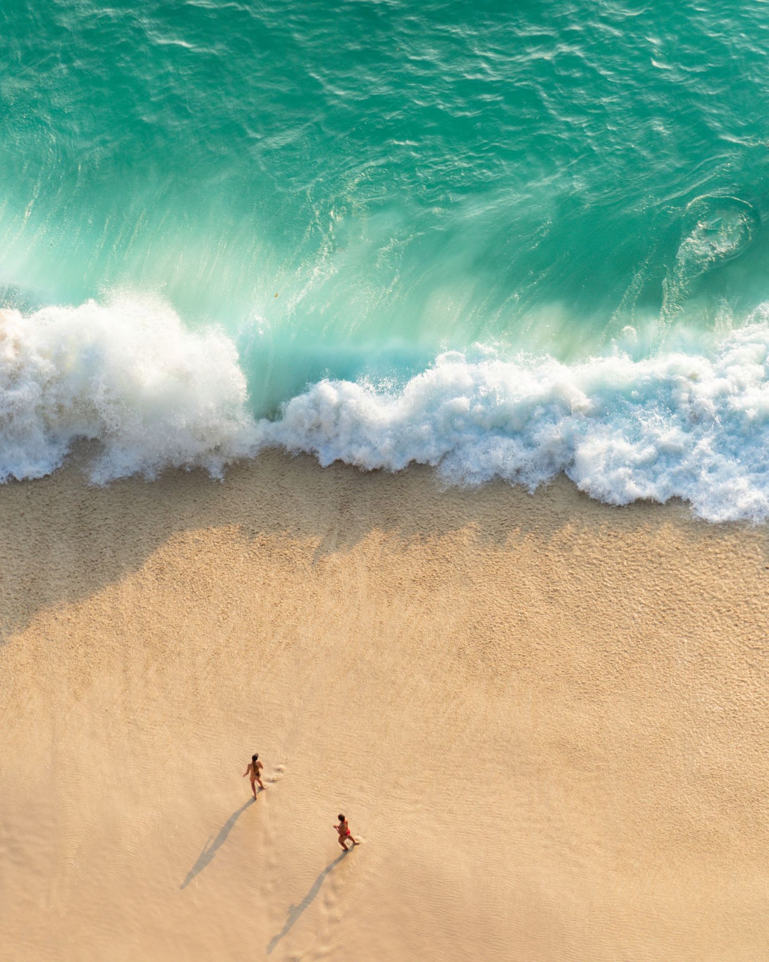 Aerial view of turquoise ocean waves meeting a pristine sandy beach with small figures walking.