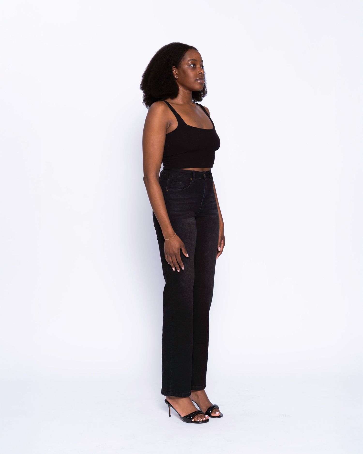 Woman in black crop top, high-waist straight-leg jeans, and heeled sandals poses sideways against white background.