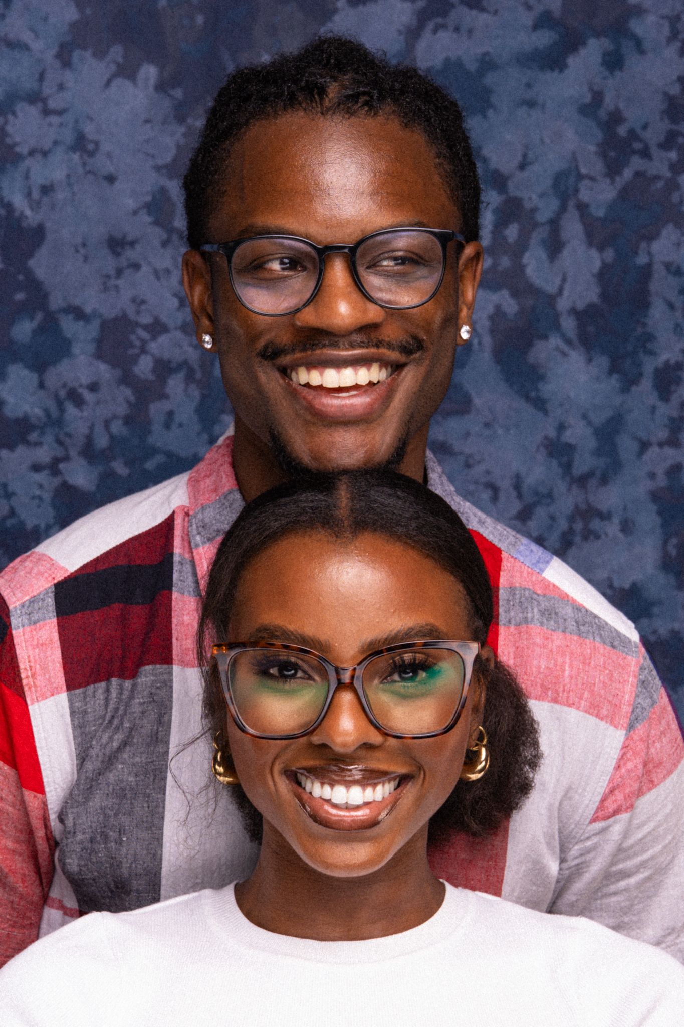 Two smiling young Black adults wearing glasses pose together against a blue mottled background.