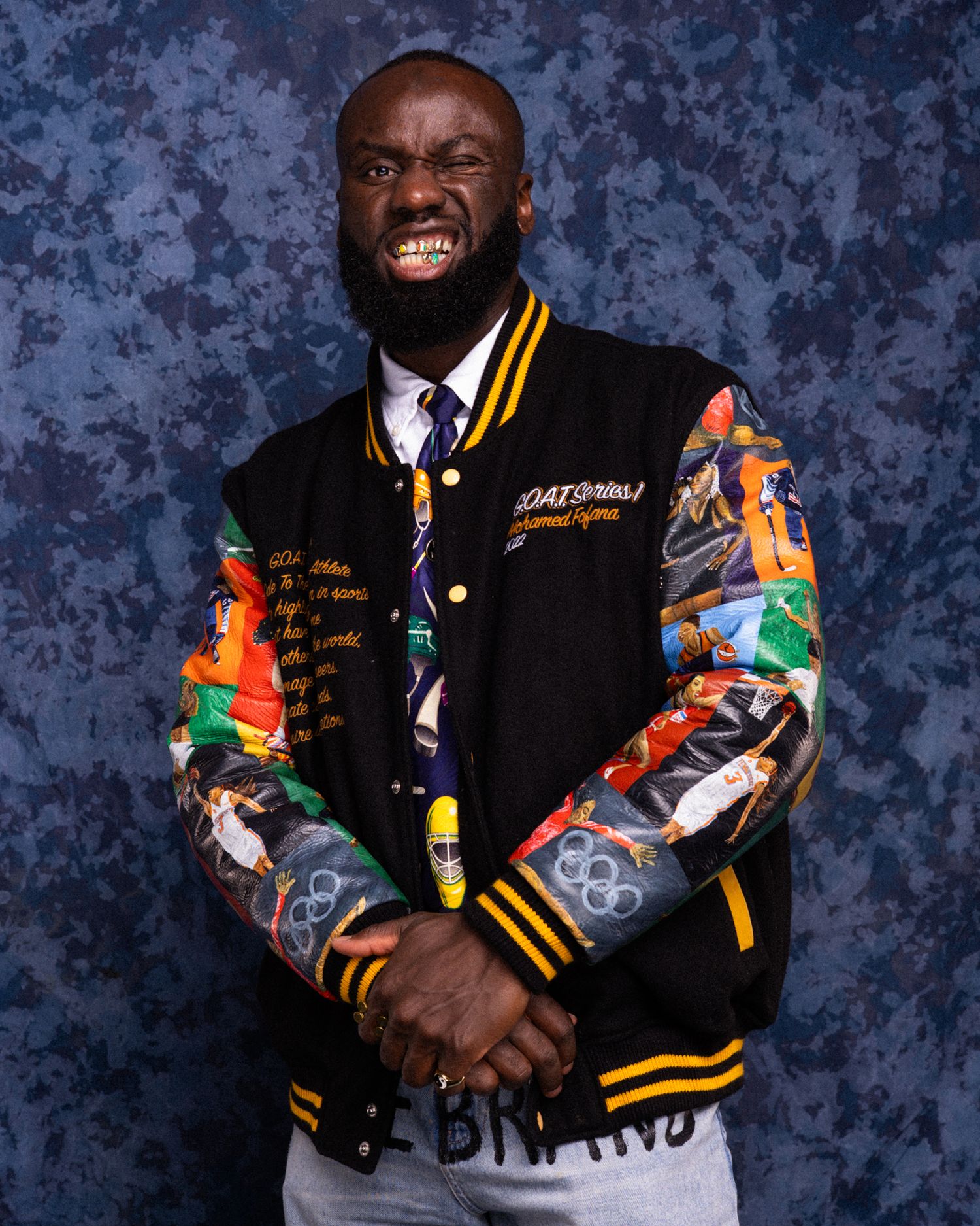 Man smiling in colorful varsity jacket with graphic sleeves, black collar, and gold trim, posing against blue background.