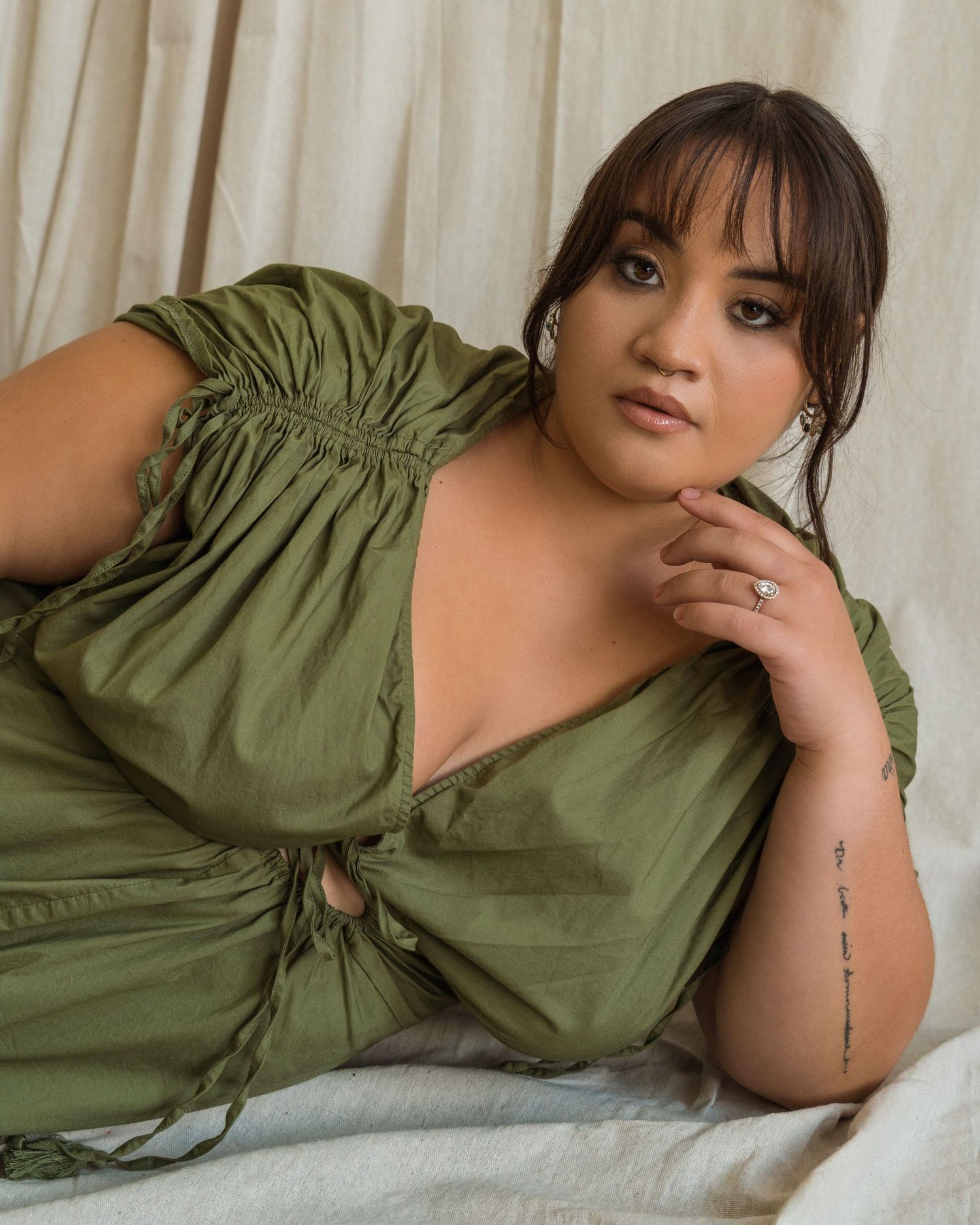 Model in olive green dress poses elegantly on white bedding while showing delicate arm tattoo.