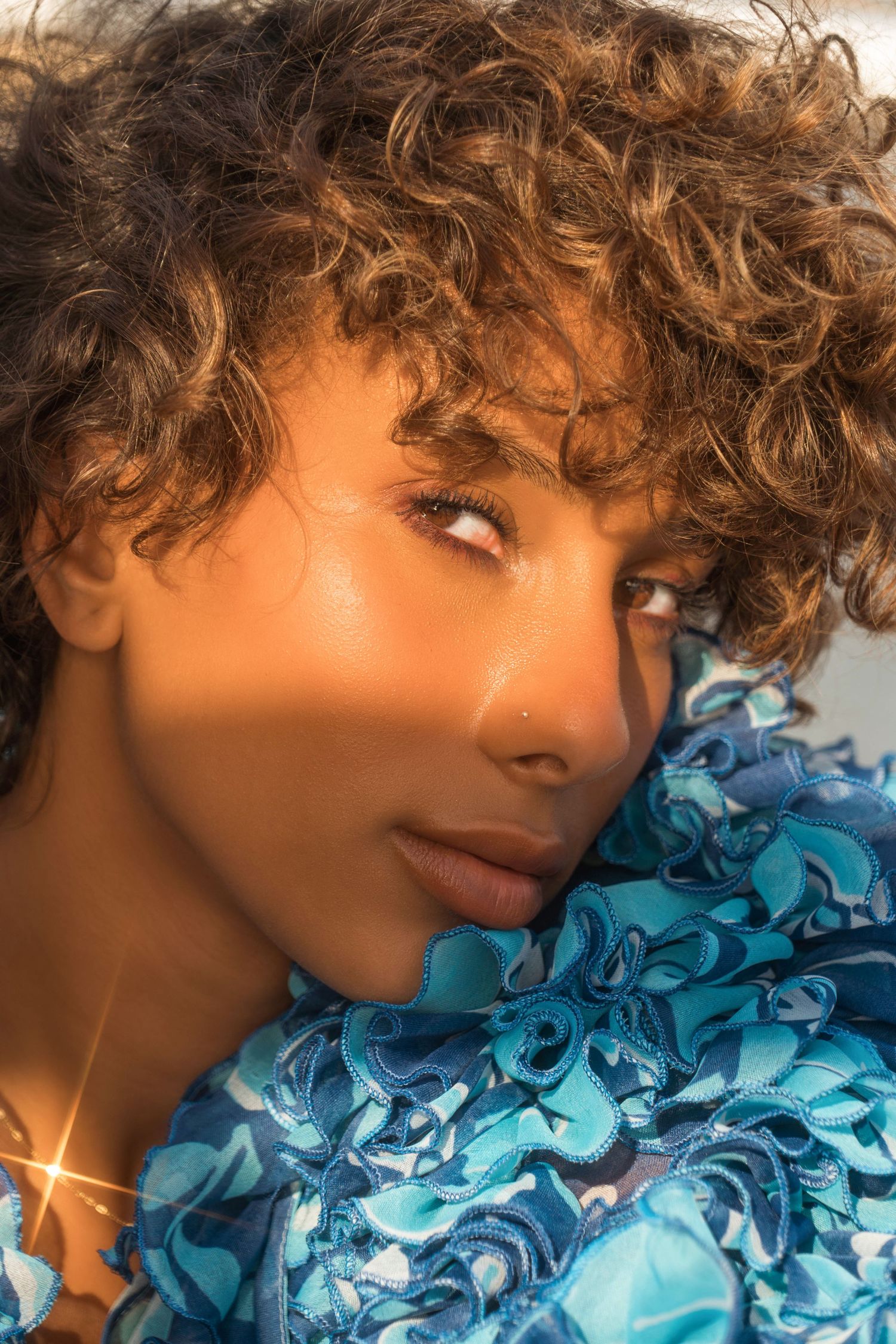 Fashion portrait with curly hair and blue ruffled fabric against warm lighting.