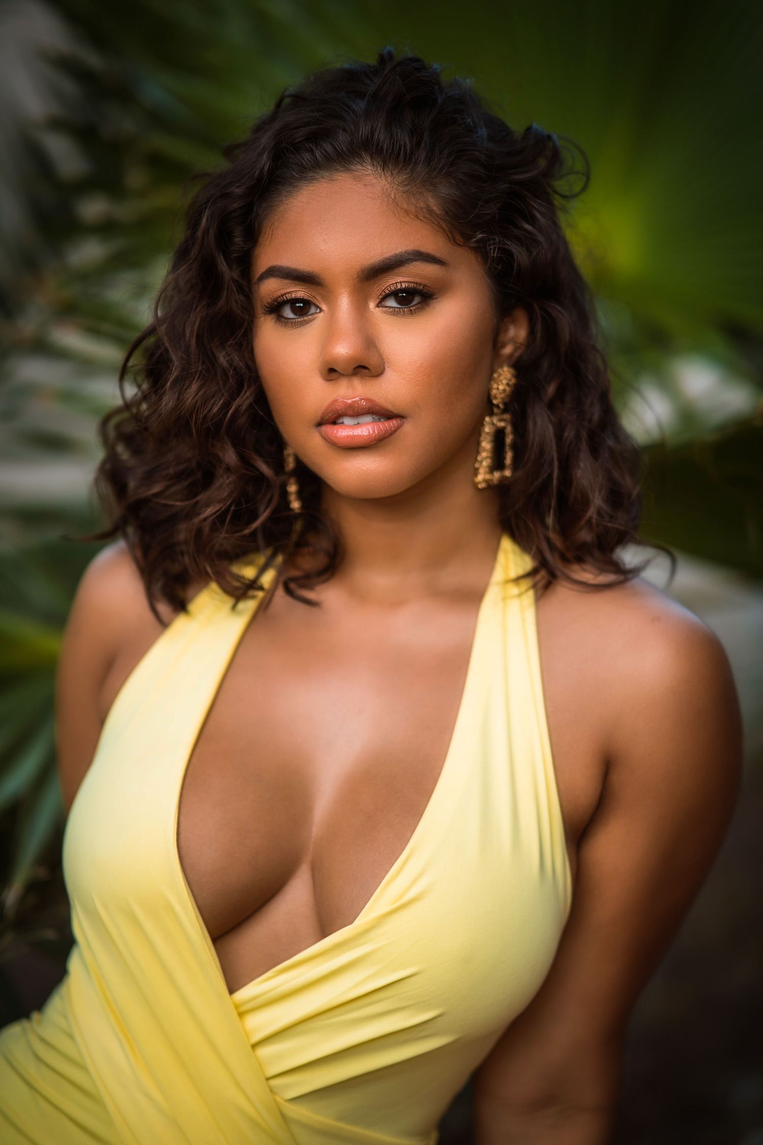 Young woman in yellow halter dress with gold earrings posing outdoors among tropical green palm leaves.