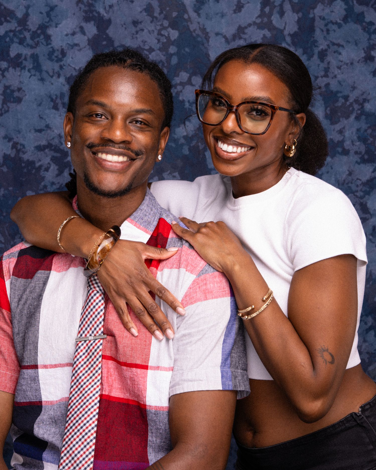 A smiling young couple poses together, the man in a plaid shirt and tie, the woman in a white crop top wearing glasses.