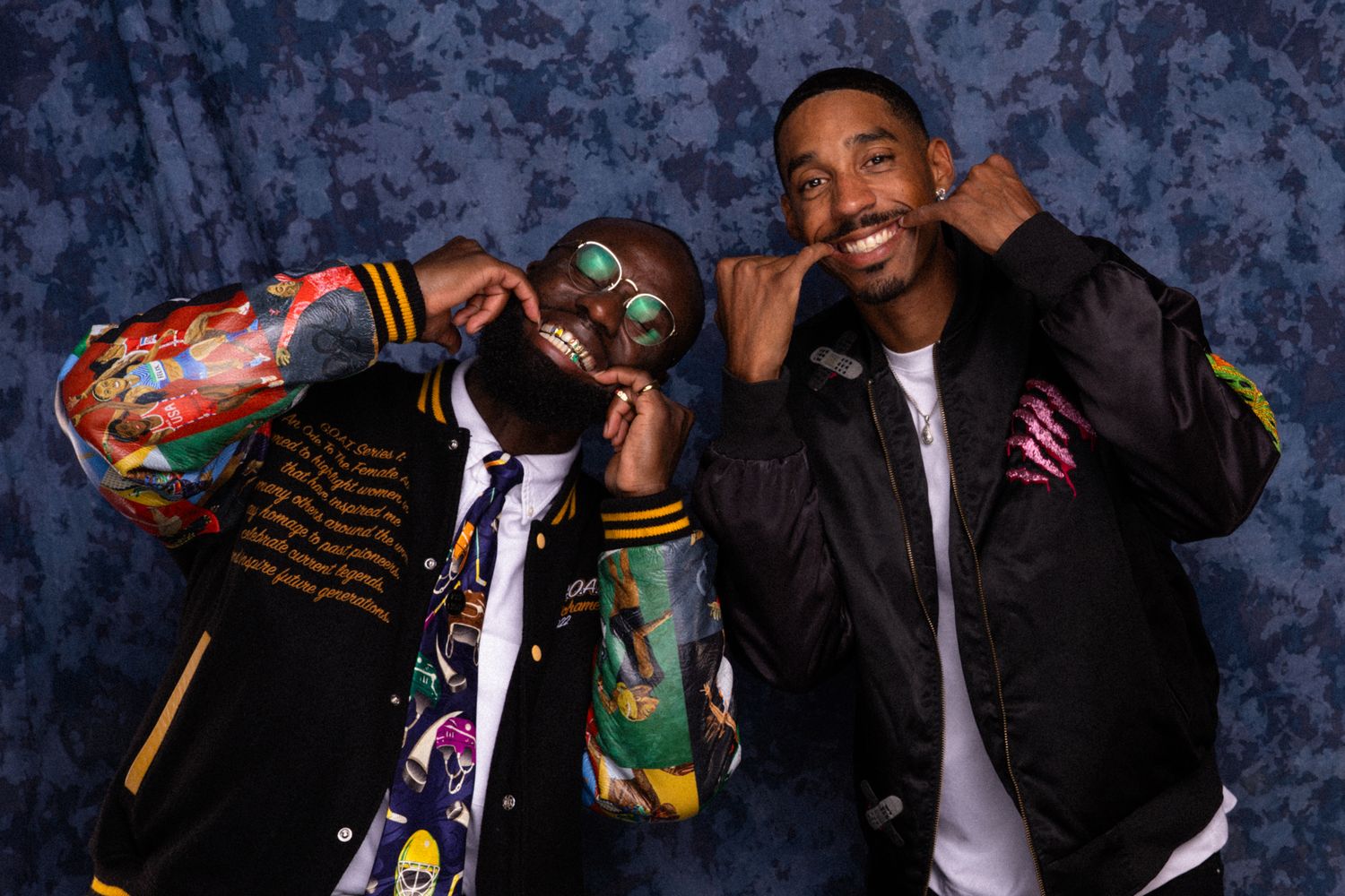 Two men posing playfully in colorful varsity jackets against a blue backdrop at a photo event.