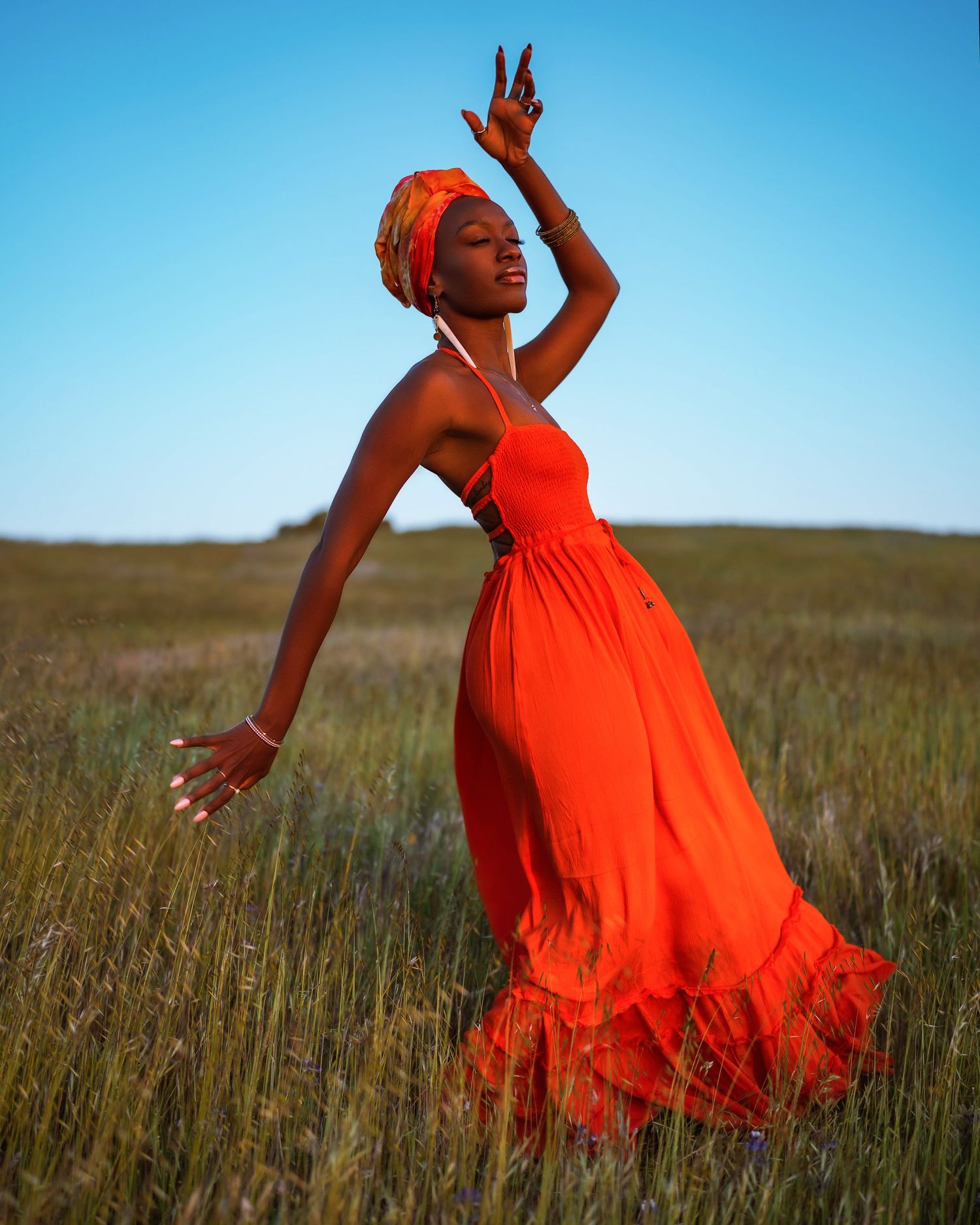 Dancer in bright orange dress twirling through tall grass against blue sky.