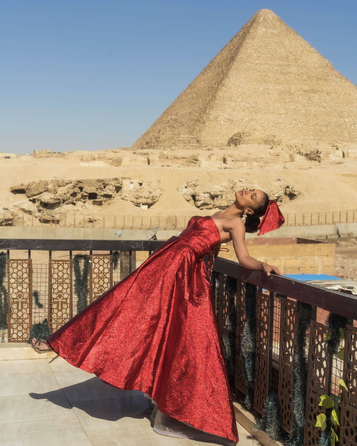 Person in red gown poses dramatically against Egyptian pyramid backdrop.