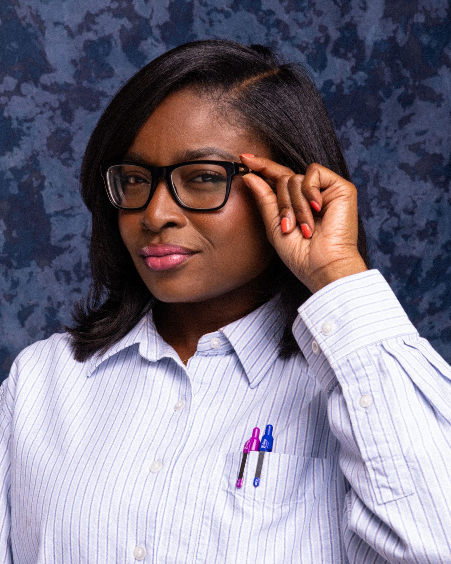 Professional woman in glasses and striped shirt with pens in pocket poses confidently against blue backdrop.