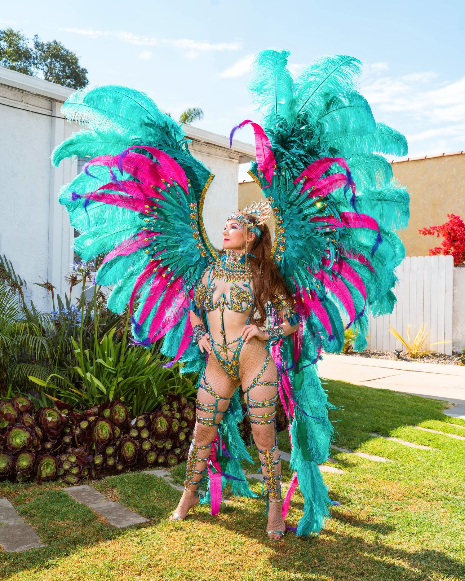 Carnival dancer in vibrant turquoise and pink feathered costume posing in backyard setting.