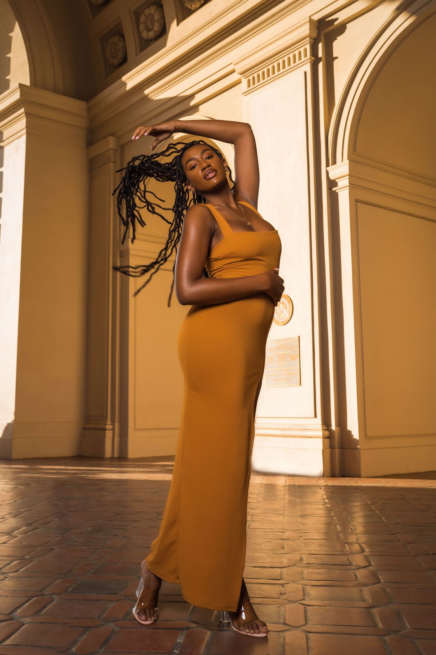 Person in mustard yellow dress poses dramatically in sunlit architectural corridor.