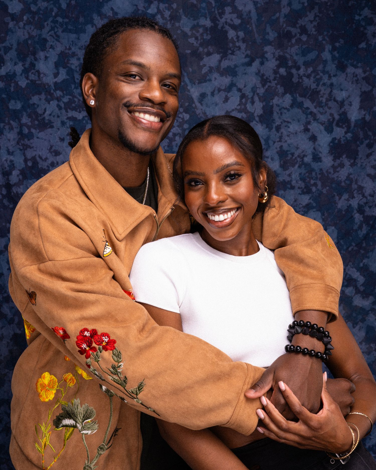 A smiling young couple poses together, man in brown floral jacket embracing woman in white top against blue backdrop.