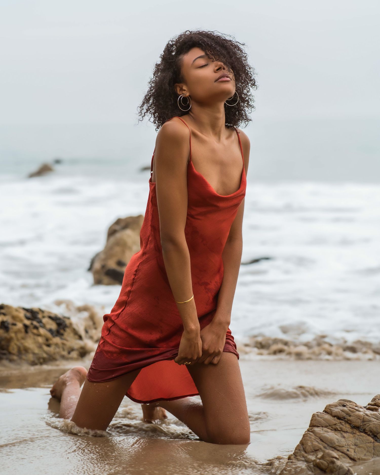 Woman in red slip dress kneeling on wet beach sand with ocean waves and rocks in background.