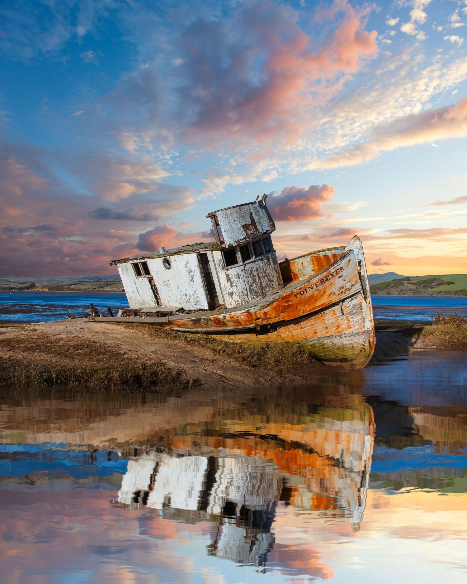 Abandoned shipwreck on beach at sunset with vibrant reflections in tidal waters.