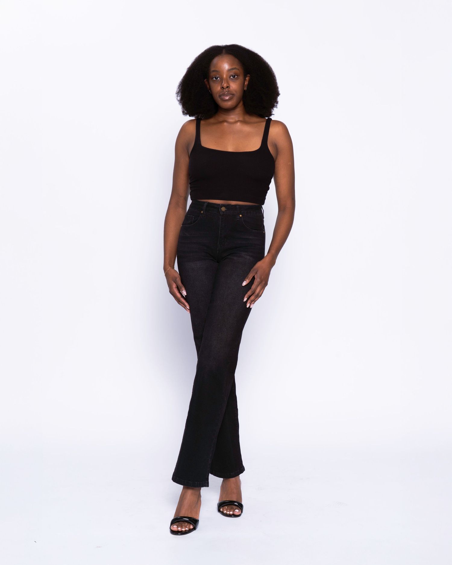 Woman wearing black crop top and high-waisted black flare jeans with black heeled sandals on white background.
