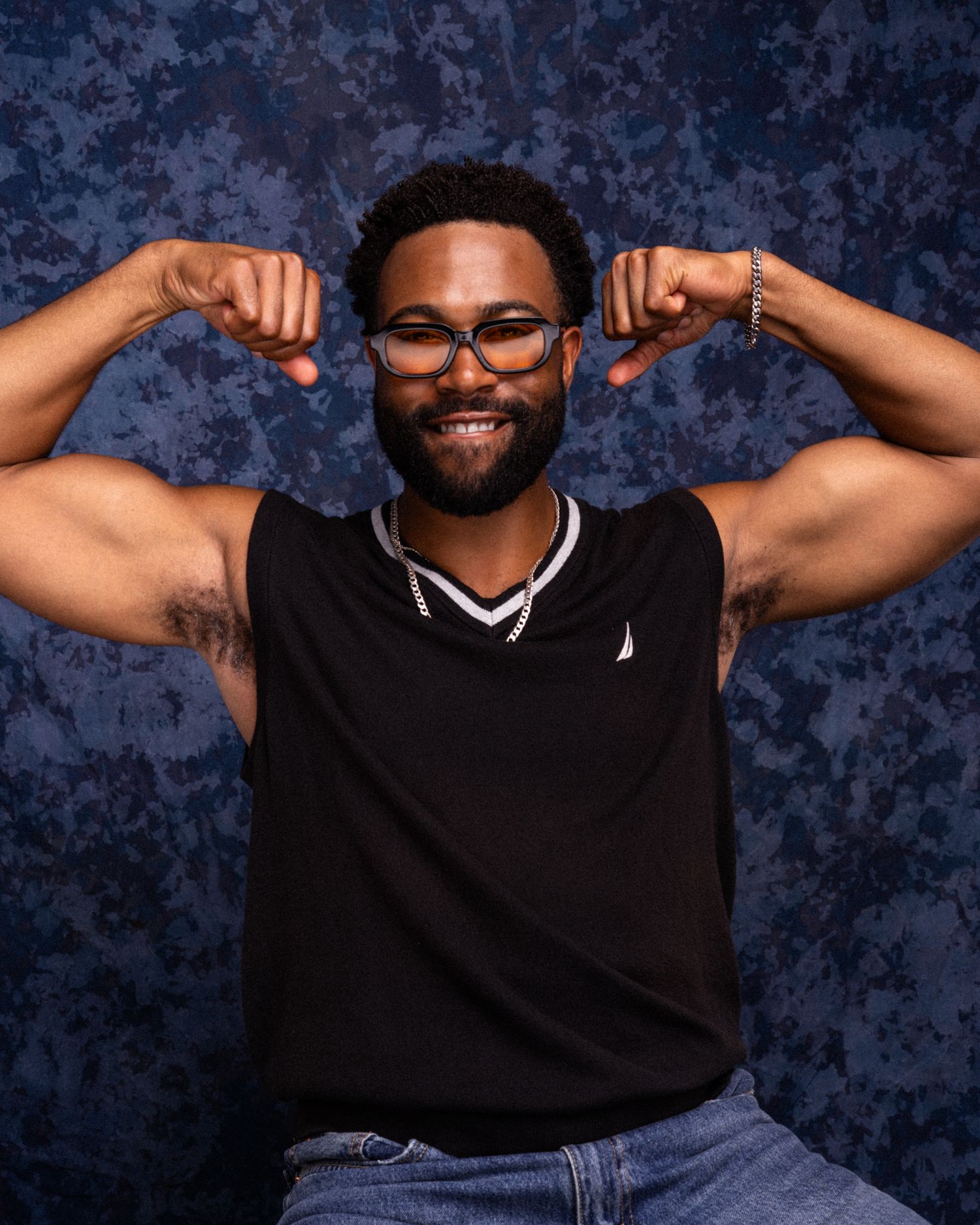 Muscular man in black Nautica tank top flexing biceps, smiling, wearing glasses and jeans.